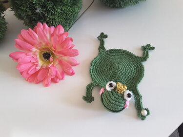 Gehäkelter grüner Frosch-Untersetzer mit Perlenakzent neben einer rosa Blume