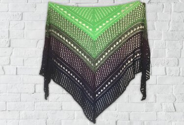 Ombre triangle crochet shawl with layered lace panels transitioning from bright green to black, hung flat on a white brick wall
