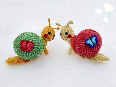 Zwei Amigurumi‑Schnecken mit spiralförmigen Schalen, applizierter Blüte und Schmetterling