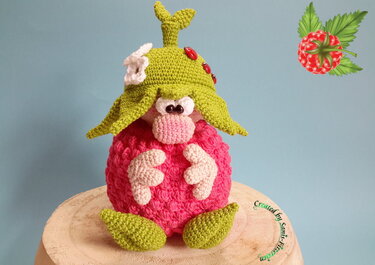 Gehäkelter Amigurumi-Wichtel in Himbeerform mit grünem Hut, weißen Blüten und roten Marienkäfern