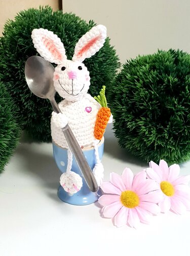 Weiß gehäkelter Osterhase als Eierwärmer in blauem Eierbecher, hält einen Löffel und eine kleine Karotte, vor grüner Deko und rosa Blumen.