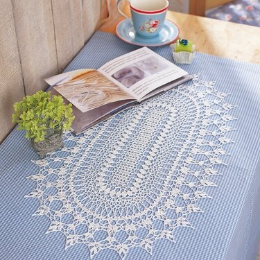 Ovales weißes gehäkeltes Deckchen mit Spitzenmuster auf blau-kariertem Tischläufer, daneben Tasse, Untertasse, offenes Buch und kleine Pflanze