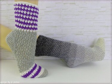 Zwei gehäkelte Socken: eine mit lila-weißem Hahnentritt am Schaft, eine mit grauem Farbverlauf.