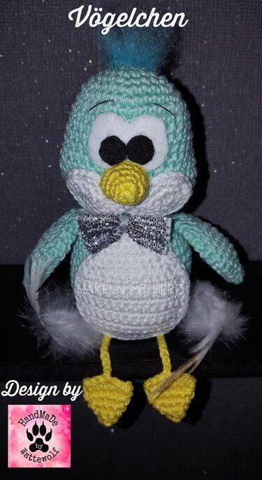 Gehäkelter Amigurumi-Vogel hellblau und weiß sitzend mit gelbem Schnabel, gelben Füßchen, schwarzem Augenpaar, blauem Schopf und silberner Glitzerfliege