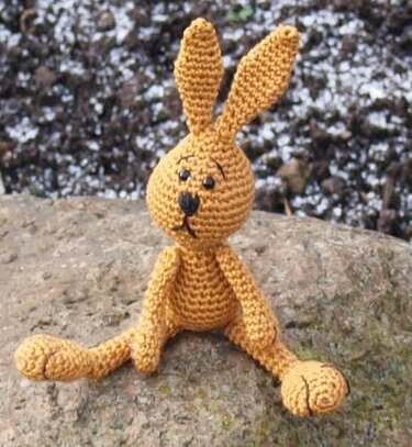 Gehäkelter senfgelber Amigurumi-Hase mit langen Ohren, sitzt auf einem Stein
