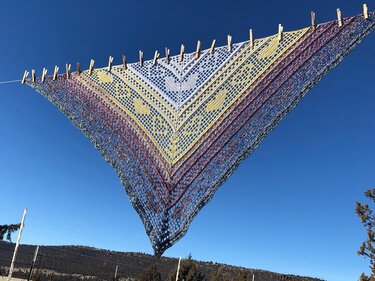 Triangular crocheted shawl with white, yellow, red and blue gradient and heart lace motifs, clipped to a clothesline against a clear blue sky.