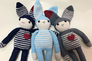 Drei gehäkelte Amigurumi-Hasen in Dunkelblau, Hellblau und Grau mit weißen Streifen und roten Herzen.