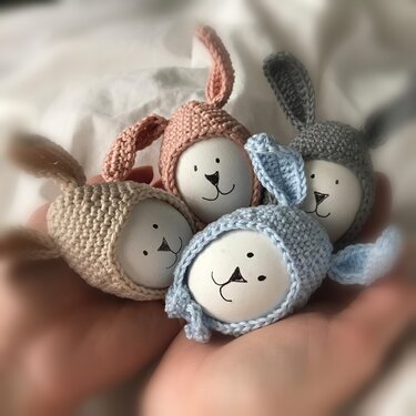 Vier gehäkelte Hasen-Eierwärmer in Pastellfarben, jeweils über bemalten Eiern, in offenen Händen liegend.
