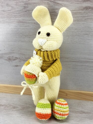 Cremefarbener Amigurumi-Hase mit weißer Schnauze und braunen Augen, trägt gelben Rollkragenpullover und hält ein gestreiftes Häkel-Ei mit Bommel; zwei weitere Häkel-Eier liegen davor.