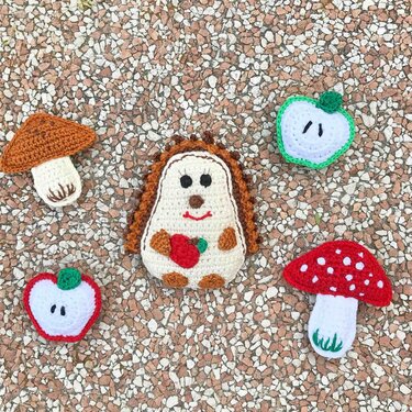 Gehäkelter Igel mit Apfel, zwei gehäkelte Pilze und zwei Apfelscheiben auf kiesigem Untergrund