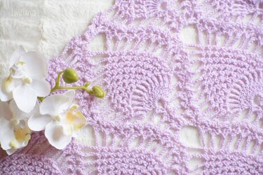 Nahaufnahme eines fliederfarbenen Häkelmusters mit weißen Orchideen auf hellem Untergrund.
