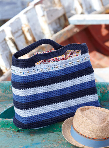 Gehäkelte Henkeltasche mit blauen und weißen Streifen, dekorative Perlen und Nieten am oberen Rand, daneben Strohhut.