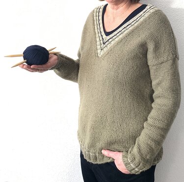Überschnittener V‑Pullover in Oliv, glatt gestrickt, mit Rippenbündchen; Garnknäuel und Stricknadeln in der Hand