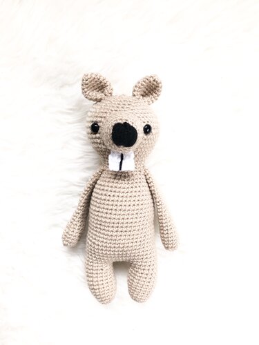 Beiger gehäkelter Amigurumi-Biber mit schwarzen Augen und zwei weißen Vorderzähnen, liegend auf hellem Untergrund
