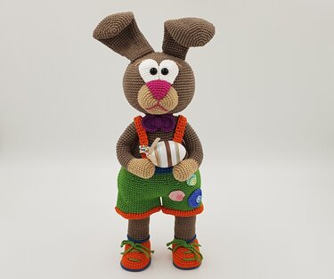 Brown crocheted amigurumi rabbit wearing green shorts, orange suspenders and a purple bow tie, holding a striped egg.