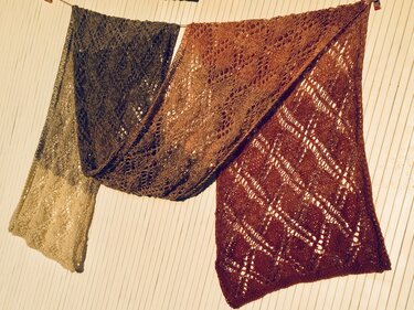 Nahansicht mehrerer gestrickter Lacetücher mit unterschiedlichen Ajour- und Blattmustern an einer Leine vor Holzpaneelen