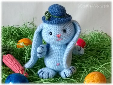 Hellblauer gehäkelter Hase mit blauem Hut, Ostergras, bunten Eiern und Häkelkarotte