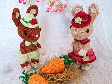Zwei gehäkelte Amigurumi-Hasen mit Hüten neben zwei gehäkelten Möhren im Nest