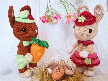 Zwei gehäkelte Amigurumi-Hasen mit Hüten, einer hält eine Möhre; davor ein Nest mit Ostereiern
