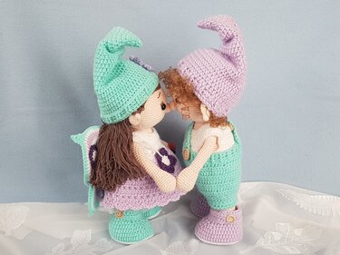 Zwei gehäkelte Amigurumi-Puppen in Mint und Lila mit Zipfelmützen und Flügeln, die sich an den Händen halten
