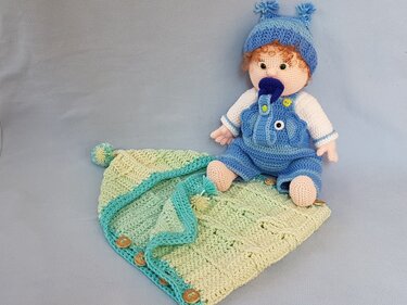 Gehäkelte Babypuppe mit blauem Strampler und Mütze auf einer hellgrün-gelben Häkeldecke mit türkiser Umrandung und Holzknöpfen.