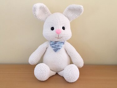 Weißer gehäkelter Amigurumi-Hase mit rosa Nase, schwarzen Augen und blau-weiß gestreiftem Halstuch auf Holzuntergrund.