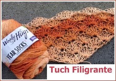 Nahaufnahme eines gehäkelten Tuchs mit filigranem Fächermuster und einem Garnknäuel in Beige- und Orangetönen auf grauem Untergrund.