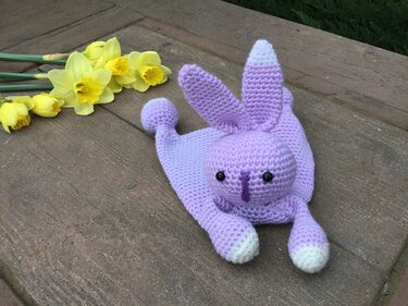 Lavender crocheted bunny snuggle blanket with white-tipped ears beside yellow daffodils on a wooden surface.
