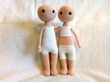 Zwei gehäkelte Amigurumi-Puppenrohlinge in Hautfarbe mit weißer Unterkleidung auf hellem Tuch.
