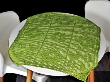 Grünes gehäkeltes Deckchen mit quadratischen Herz- und Blumenmotiven auf rundem weißen Tisch