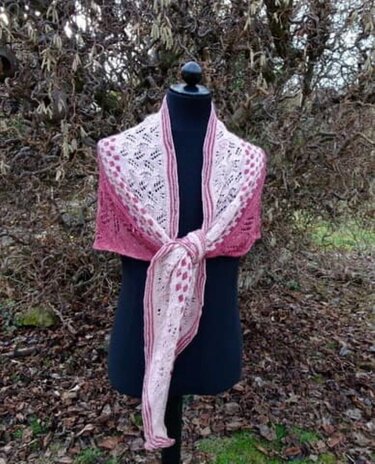 Gestricktes Dreieckstuch in Rosa und Creme mit Lochmuster, drapiert auf einer schwarzen Schneiderpuppe vor Sträuchern.