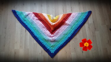 Dreieckstuch gehäkelt mit regelmäßigen GrannyReihen und Farbverlauf, liegt flach auf Holzfußboden neben einer roten Blume