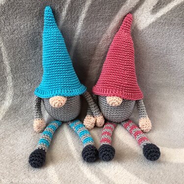 Zwei gehäkelte Amigurumi-Wichtel mit spitzen Hüten (blau und pink) auf grauer Decke