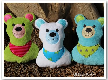 Drei gestickte Kuschelbären in Grün, Weiß und Hellblau mit Halstüchern vor einem Holzstamm auf Nadelholzmulch.