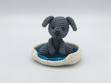 Gehäkelter grauer Amigurumi‑Hund im runden weiß‑blauen Körbchen