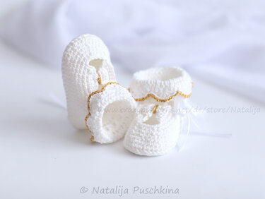 Zwei weiße gehäkelte Babyschuhe mit goldener Borte und kleinen Schleifen auf hellem Hintergrund.
