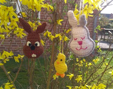 Drei gehäkelte Osteranhänger (brauner Hase, heller Hase, gelbes Küken) hängen an einem Forsythienzweig mit gelben Blüten
