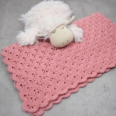 Pink crocheted baby blanket with scalloped edge and shell stitch pattern, styled with a plush lamb toy
