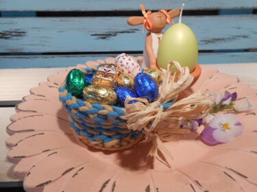 Gehäkeltes blau-beiges Osternest mit Schokoeiern und Raffia-Schleife auf rosa Platte