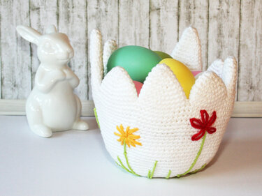 Weißer, gehäkelter Osterkorb in Tulpenform mit bunten Ostereiern und bestickten Blumen; keramischer Hase daneben