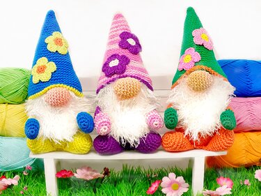 Three crocheted gnomes with flower-decorated pointed hats and fluffy white beards seated on a white bench with colorful yarn in the background.