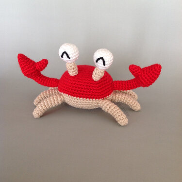 Red and beige crocheted amigurumi crab toy with white crocheted eyes, on a grey background.