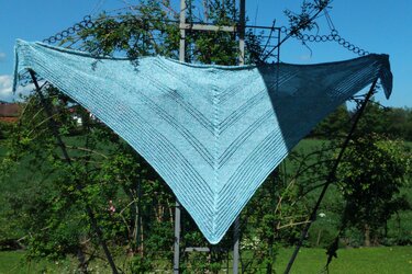 Light blue knitted triangle shawl with V-shaped garter and eyelet pattern, draped on a garden trellis.