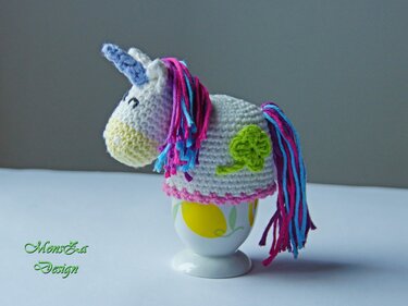 Gehäkelter weißer Einhorn-Eierwärmer mit rosa-blauer Mähne und grünem Kleeblatt auf Eierbecher