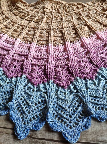 Nahaufnahme eines gehäkelten Halbrundtuchs mit Farbverlauf von Beige über Rosa nach Blau, filigranem Lochmuster, vertikalen Säulen und einem Zackenrand.