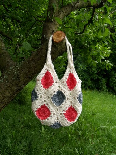 Gehäkelte Henkeltasche aus Granny Squares in Creme mit roten und grauen Mittelpunkten hängt an einem Baum