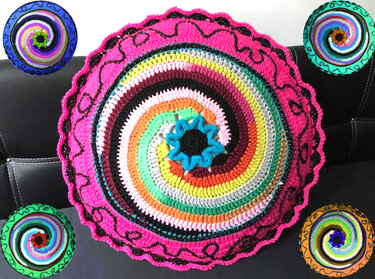 Rundes gehäkeltes Kissen mit buntem Spiralmuster und pinker Umrandung auf dunklem Sofa.