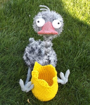 Gehäkelter Amigurumi-Vogel grau mit rosafarbenem Schnabel und gelbem Korb auf grünem Gras