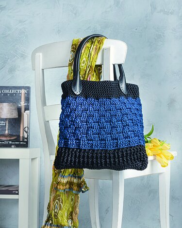 Gehäkelte Henkeltasche in Blau und Schwarz mit Strukturmuster, auf einem weißen Stuhl