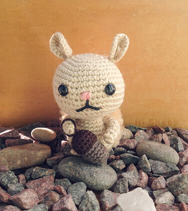 Beige crocheted amigurumi bunny with dark round eyes and pink nose holding a brown crocheted acorn, seated on gray and reddish rocks.